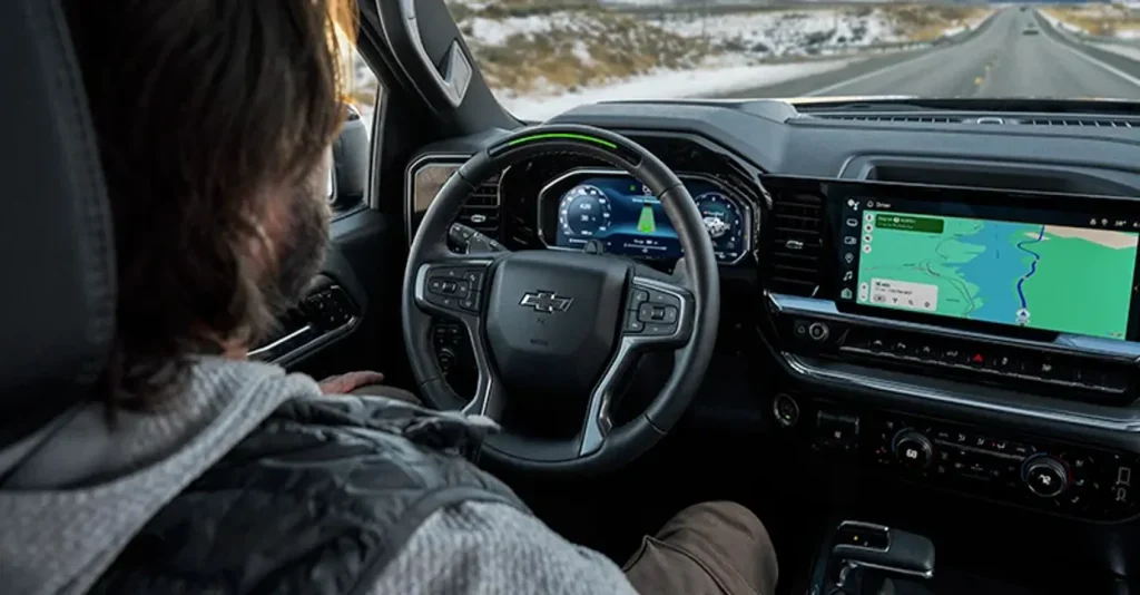The image shows a person driving a Chevrolet Silverado, with a clear view of the advanced digital dashboard and navigation system. The snowy road and mountainous scenery outside highlight the vehicle's rugged, yet tech-savvy design.