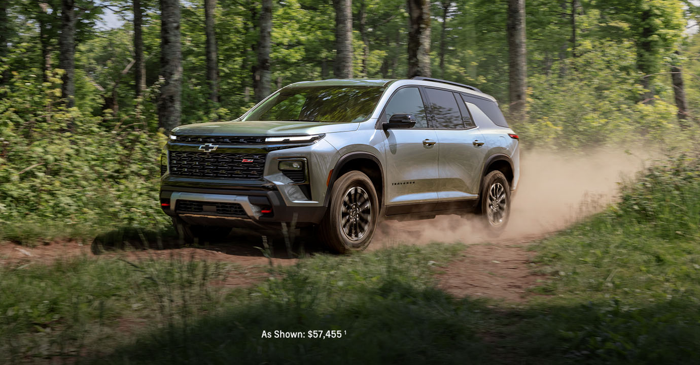 A 2026 Chevrolet Traverse SUV driving on a dirt trail through a wooded forest, kicking up dust as it moves along the rugged path.