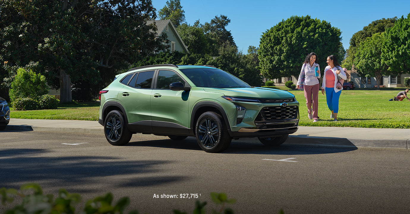 2026 Chevy Trax green compact SUV parked on a suburban street with two women walking nearby in a sunny neighborhood setting