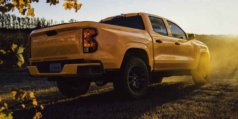 A yellow pickup Chevrolet Colorado ZR2 Bison on a dirt road at sunset, surrounded by autumn leaves. The scene conveys ruggedness and adventure with a warm, golden glow.