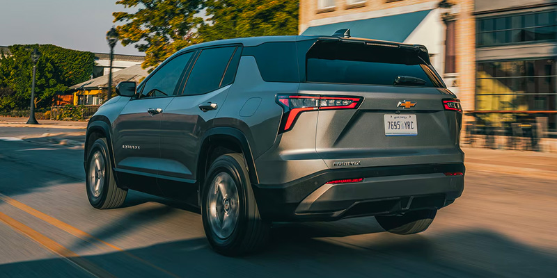 A silver Chevrolet Equinox drives on a sunlit road beside buildings and trees. The vehicle's rear and taillights are visible, conveying motion and modern design.